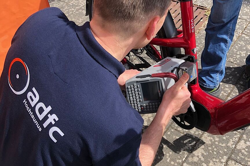 Ein Fahrrad wird codiert Man sieht einen Mann im ADFC-Hemd vor einem Fahrrad knien, mit einem Codiergerät, das er an der Sattelstange des vor ihm stehenden Fahrrads ansetzt. Die Szene spielt sich am Stand des ADFC OG Kronberg auf dem bunten Flohmarkt der Altkönigschule am 25.5.2025 ab.