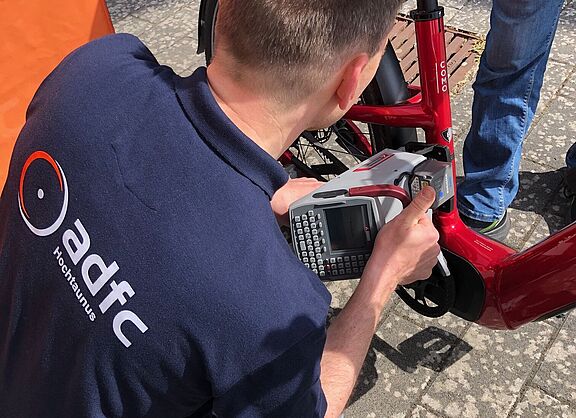 Man sieht einen Mann im ADFC-Hemd vor einem Fahrrad knien, mit einem Codiergerät, das er an der Sattelstange des vor ihm stehenden Fahrrads ansetzt. Die Szene spielt sich am Stand des ADFC OG Kronberg auf dem bunten Flohmarkt der Altkönigschule am 25.5.2025 ab.