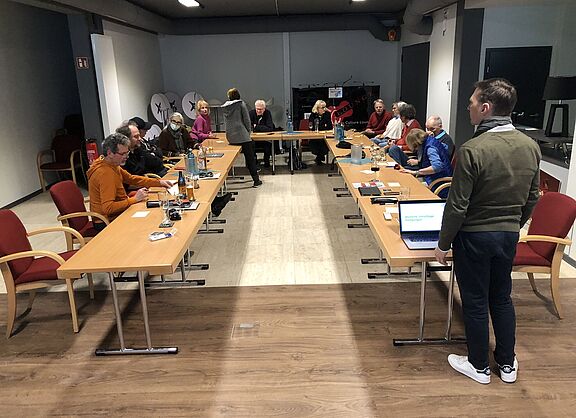 Ein Bild von der Jahresversammlung 2024 in Kronberg. Ein Mann steht vor einer U-Formation von Tischen, an welchen die Mitglieder sitzen.