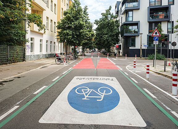 Eine Straße in Berlin: Rechts und links sind Häuserzeilen zu sehen, die Fahrbahn ist mit einem großen Fahrradsymbol versehen. Weiter hinten ist die Fahrbahn rot eingefärbt, Poller dienen als modaler Filter.  